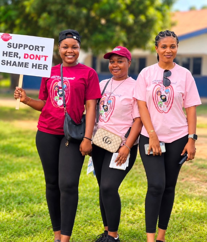 Three volunteers in summit T-shirts, one holding a placard reading Support her, don't shame her
